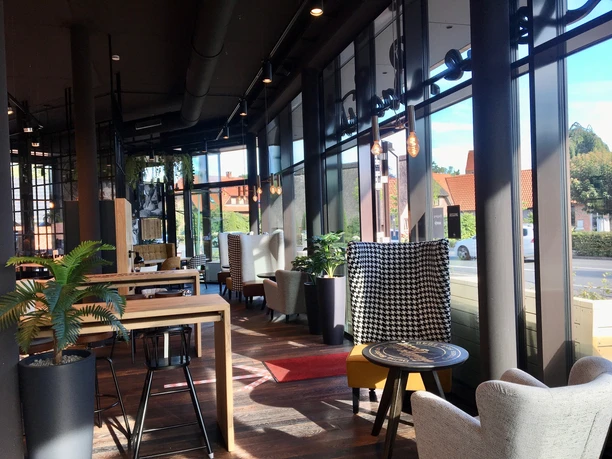 Müller & Egerer Ein gemütliches Café mit hohen Fenstern, stilvollen Polstermöbeln und viel natürlichem Licht. Pflanzen stehen in modernen Töpfen und Holzmöbel verleihen dem Raum Wärme.