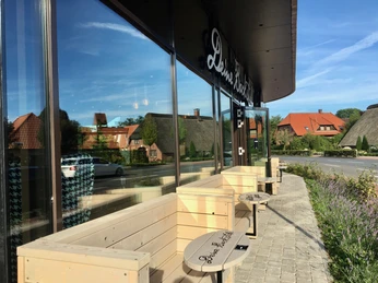 Müller & Egerer Das Bild zeigt eine moderne Café-Terrasse von Müller & Egerer mit gemütlichen Holzbänken und Tischen vor einem gläsernen Gebäude. Im Hintergrund sind traditionelle Reetdachhäuser zu sehen, eingerahmt von einem klaren blauen Himmel.