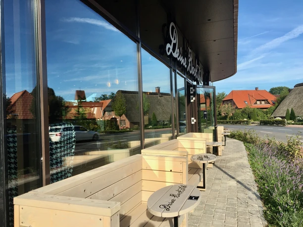 Müller & Egerer Das Bild zeigt eine moderne Café-Terrasse von Müller & Egerer mit gemütlichen Holzbänken und Tischen vor einem gläsernen Gebäude. Im Hintergrund sind traditionelle Reetdachhäuser zu sehen, eingerahmt von einem klaren blauen Himmel.