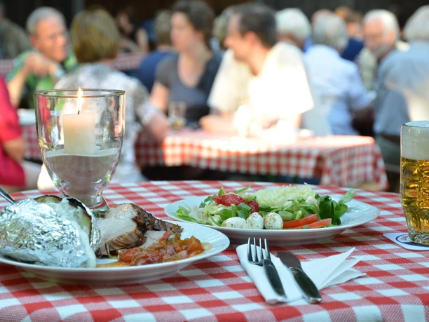 Heidjertypische Gastlichkeit Deftige Speisen, Salat und Bier auf einem Tisch mit rot-weißer Decke im Außenbereich.