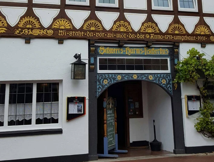 Fachwerkhaus mit traditionellen Verzierungen und Rankpflanzen am Eingang; Schild: "Schweers Harms Fischerhus".