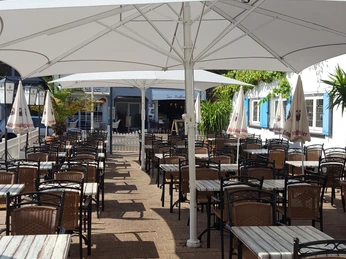 Sonniger Außenbereich eines Cafés mit leeren Tischen und Stühlen unter weißen Sonnenschirmen.