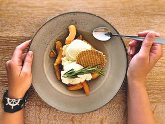 Vanille Eis Ein Teller mit Vanilleeis, Apfelschnitzen, einem Waffelblatt und Rosmarinzweig, von Händen gehalten.