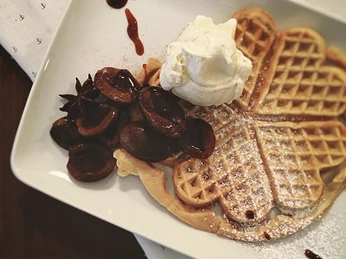 Herzförmige Waffel mit Puderzucker, Schlagsahne und saftigen Pflaumenstücken auf einem weißen Teller.