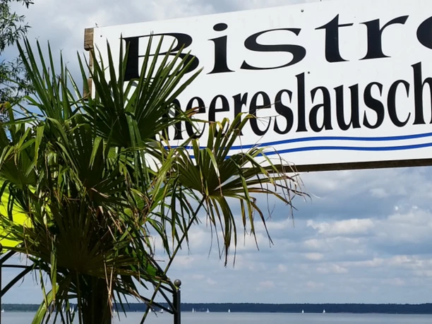 Bistro Meereslauschen Bistro Meereslauschen: Schild vor Palmen und Blick auf den See mit blauem Himmel und Wolken.
