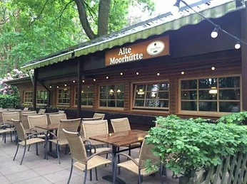Alte Moorhütte Ein traditionelles Holzgebäude mit Tischen und Stühlen auf der Terrasse umgeben von üppigem Grün.