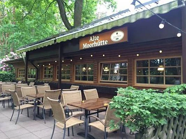 Alte Moorhütte Ein traditionelles Holzgebäude mit Tischen und Stühlen auf der Terrasse umgeben von üppigem Grün.
