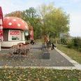 Café mit Fliegenpilz-Dach in herbstlicher Umgebung, umgeben von buntem Laub und Sitzmöglichkeiten.Café with toadstool roof in autumnal surroundings, surrounded by colorful foliage and seating.Café med paddehattetag i efterårsagtige omgivelser, omgivet af farverige blade og siddepladser.Café met paddenstoeldak in een herfstachtige omgeving, omringd door kleurrijk gebladerte en zitjes.