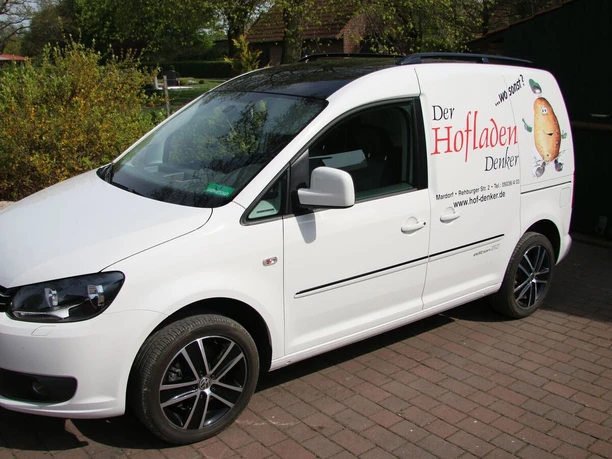 Hofladen Denker Ein weißer Kleintransporter mit Hofladen-Beschriftung parkt auf einem gepflasterten Hof bei sonnigem Wetter.