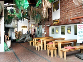 Fischerei & Aalräucherei Hodann GmbH Die Fischerei & Aalräucherei Hodann GmbH bietet rustikale Holztische unter hängenden Fischernetzen.