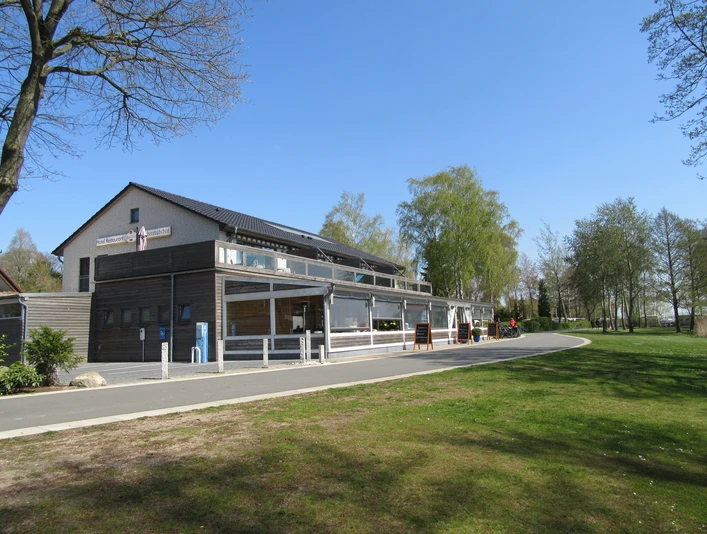 Fischerstübchen Ein modernes Restaurant mit großen Glasfronten, umgeben von Bäumen und angrenzend an eine ruhige Straße.