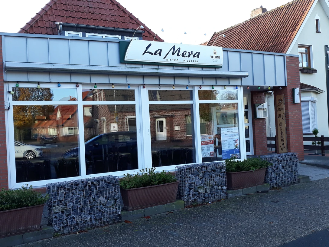 Pizzeria La Mera Rote Backsteinfassade mit weißen Fensterstreben und Schriftzug "La Mera" über der Eingangstür.