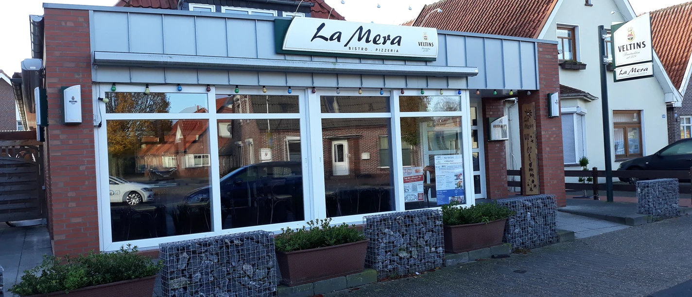 Pizzeria La Mera Rote Backsteinfassade mit weißen Fensterstreben und Schriftzug "La Mera" über der Eingangstür.
