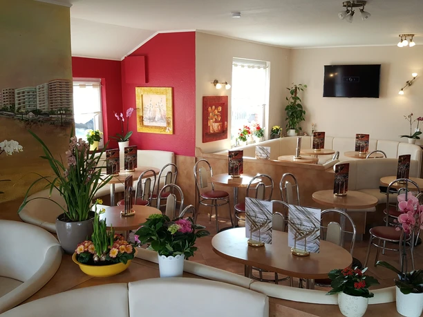 Eiscafé Venezia Innenansicht Das Bild zeigt den schicken Innenbereich des Eiscafés Venezia mit hellen Ledersitzbänken und runden Holztischen, umgeben von üppigen Pflanzen. Warme Farben und dezente Beleuchtung schaffen eine entspannte, einladende Atmosphäre.