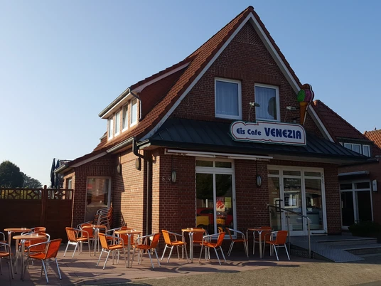Eiscafé Venezia Außenansicht Die Außenansicht des Eiscafé Venezia zeigt ein Backsteingebäude mit orangen Stühlen und Tischen auf der Terrasse bei strahlendem Wetter.