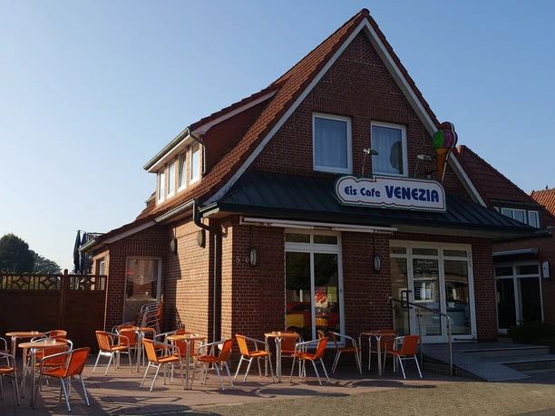 Eiscafé Venezia Außenansicht Die Außenansicht des Eiscafé Venezia zeigt ein Backsteingebäude mit orangen Stühlen und Tischen auf der Terrasse bei strahlendem Wetter.