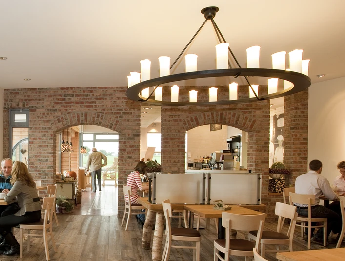 Bäckerei Ripken Innenansicht Einladendes Café mit Backsteinwänden, Holzmöbeln und großen, rustikalem Kronleuchter in Kerzenoptik. Gäste genießen das Ambiente bei Kaffee und Gebäck.Inviting café with brick walls, wooden furniture and a large, rustic candle-look chandelier. Guests enjoy the ambience with coffee and pastries.Indbydende café med murstensvægge, træmøbler og en stor, rustik lysekrone med stearinlys. Gæsterne nyder atmosfæren med kaffe og kager.Uitnodigend café met bakstenen muren, houten meubels en een grote, rustieke kroonluchter met kaarsen. Gasten genieten van de sfeer met koffie en gebak.