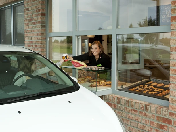 Bäckerei Ripken Drive in Ein Auto hält am Drive-in-Fenster der Bäckerei Ripken, während eine freundliche Verkäuferin Backwaren und ein Getränk überreicht.