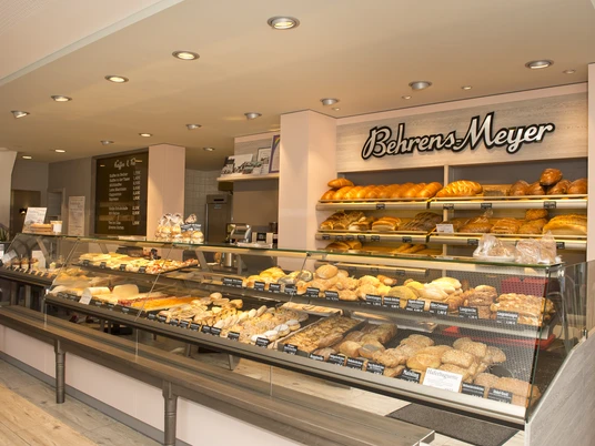 Innenansicht Cafe Behrens-Meyer Die Innenansicht des Cafés Behrens-Meyer zeigt eine einladende Theke, voll bestückt mit frischen Backwaren. An der Wand dahinter sind dekorativ das Logo des Cafés und verschiedene köstlich aussehende Brotsorten arrangiert.
