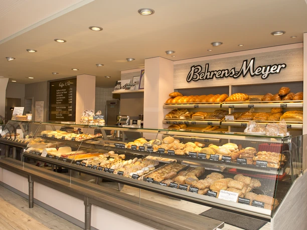 Innenansicht Cafe Behrens-Meyer Die Innenansicht des Cafés Behrens-Meyer zeigt eine einladende Theke, voll bestückt mit frischen Backwaren. An der Wand dahinter sind dekorativ das Logo des Cafés und verschiedene köstlich aussehende Brotsorten arrangiert.
