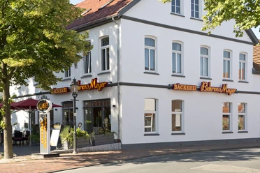 Aussenansicht Cafe Behrens-Meyer Das Bild zeigt die Außenansicht des Cafés Behrens-Meyer, ein charmantes weißes Gebäude mit großen Fenstern, umgeben von Bäumen und einer einladenden Terrasse mit Sonnenschirmen. Leuchtende Schilder weisen auf die Bäckerei hin.