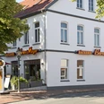 Aussenansicht Cafe Behrens-Meyer Das Bild zeigt die Außenansicht des Cafés Behrens-Meyer, ein charmantes weißes Gebäude mit großen Fenstern, umgeben von Bäumen und einer einladenden Terrasse mit Sonnenschirmen. Leuchtende Schilder weisen auf die Bäckerei hin.