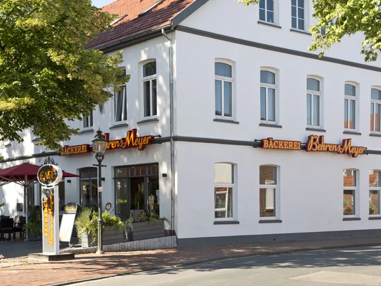 Aussenansicht Cafe Behrens-Meyer Das Bild zeigt die Außenansicht des Cafés Behrens-Meyer, ein charmantes weißes Gebäude mit großen Fenstern, umgeben von Bäumen und einer einladenden Terrasse mit Sonnenschirmen. Leuchtende Schilder weisen auf die Bäckerei hin.