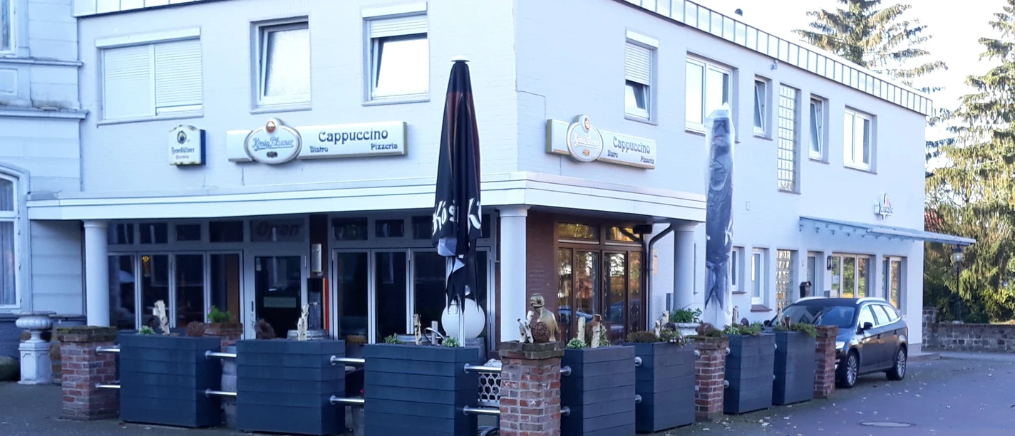 Bistro Cappuccino Das Bild zeigt das Bistro Cappuccino, einen zweistöckigen, weiß verputzten Bau mit markanten schwarzen Sonnenschirmen und modernen Außensitzbereichen, die mit Pflanzkästen geschmückt sind. Im Hintergrund parkt ein Auto. Die Schilder über den Fenstern tragen den Bistro-Namen.