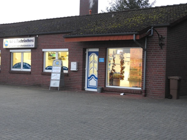 Aal- und Fischräucherei Prahm The Prahm eel and fish smokehouse is a red brick building with large shop windows through which lights stream. A blue and white sign hangs above the door and a display informs customers about the current range of products on offer. The forecourt is made of paving stones.