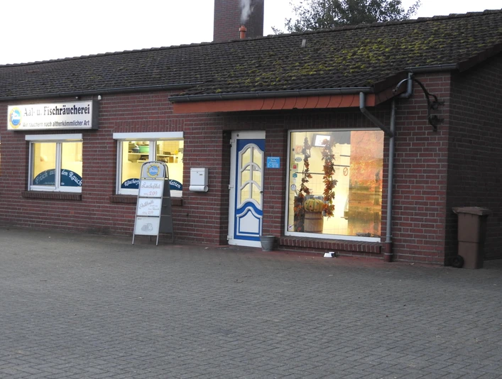 Aal- und Fischräucherei Prahm Die Aal- und Fischräucherei Prahm ist ein rotes Backsteingebäude mit großen Schaufenstern, durch die Lichter strömen. Ein blau-weißes Schild hängt über der Tür, und ein Aufsteller informiert über das aktuelle Angebot. Der Vorplatz besteht aus gepflasterten Steinen.