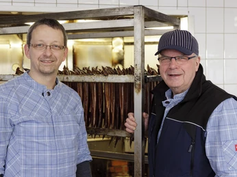 beim Räuchern Zwei Männer lächeln vor einer Räucherstation mit hängenden Fischen im Hintergrund in einem hellen Raum.