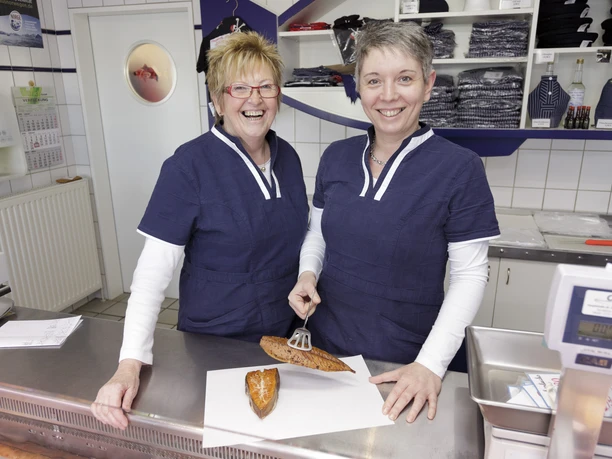 Two cheerful saleswomen in blue fishermen's shirts present smoked fish behind the counter.