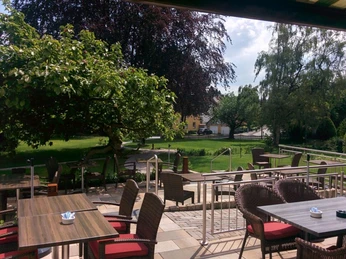 Schulze Ladencafé, Borgholzhausen Terrasse eines Cafés mit Holztischen und geflochtenen Stühlen, umgeben von Bäumen und Grünfläche.