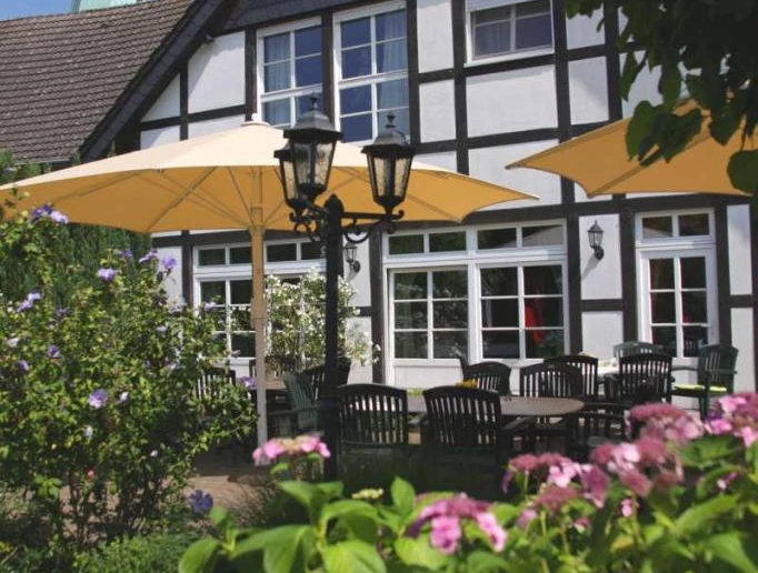 Biergarten am Café zur Linde Sonniger Biergarten mit Holztischen, blühenden Sträuchern und weißen Fachwerkhaus im Hintergrund.