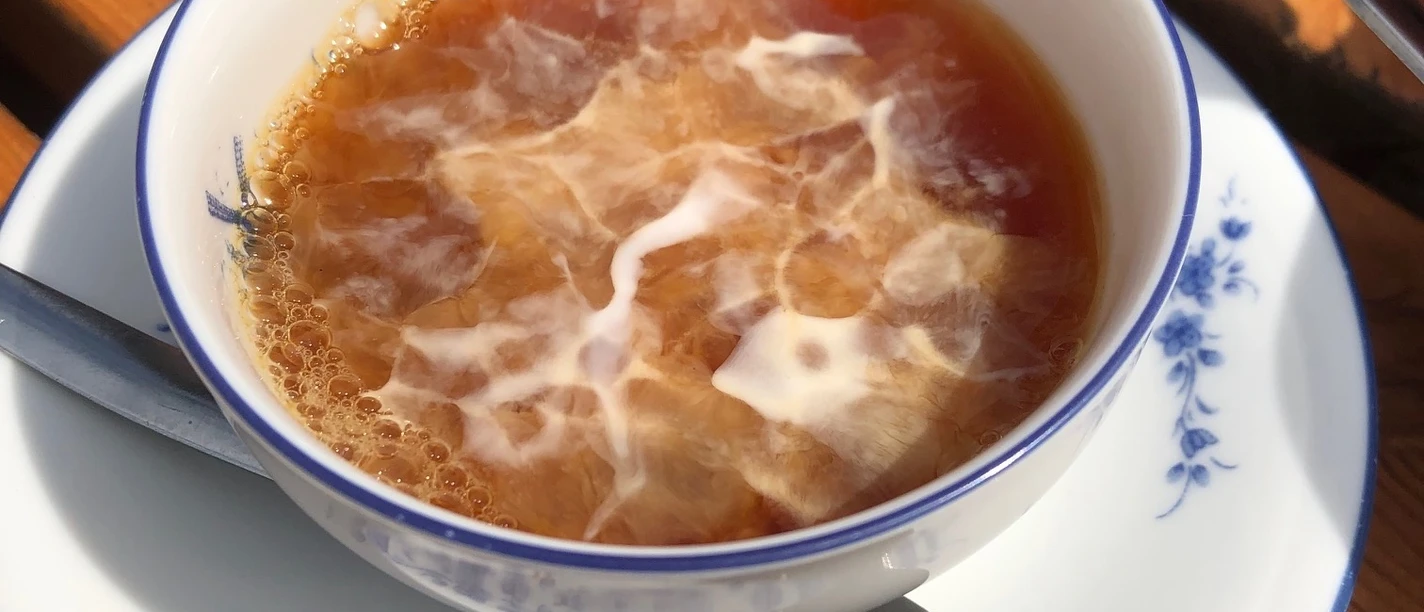Teetied_waddensea.travel_ostfriesland.jpg Ostfriesentee mit einem Sahnewölkchen in einer weiß-blauen Tasse auf einem Holztisch.