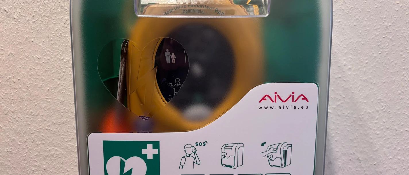 Defibrillator TI Mardorf Der Defibrillator in der Tourist-Information in Mardorf. Das orangene Gerät hängt in einem grünen Kasten vor einer weißen Wand.