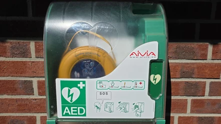 Defibrillator Wohnmobilstellplatz Steinhude Der Defibrillator am Wohnmobilstellplatz in Steinhude. Das orangene Gerät hängt in einem grünen Kasten vor einer Ziegelsteinwand.