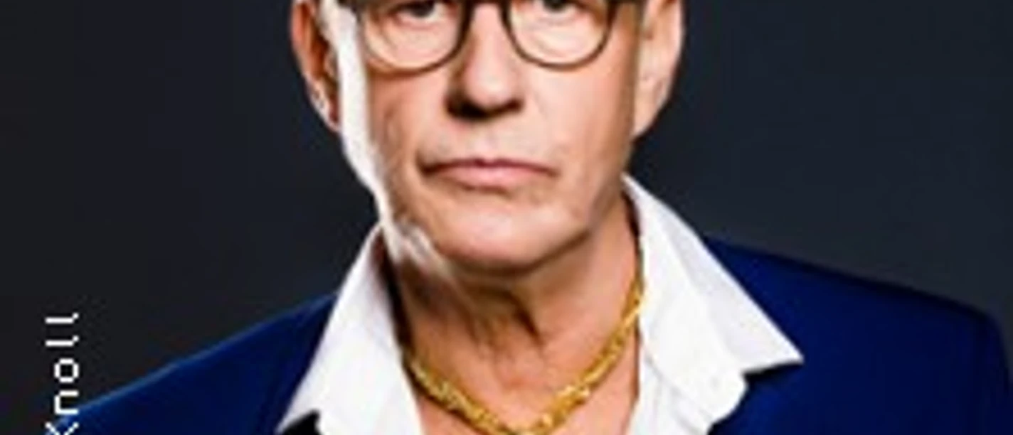 Mann mit Brille im blauen Anzug und goldener Dollar-Kette vor dunklem Hintergrund