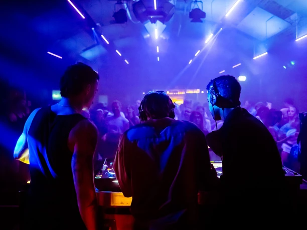 Sissy that rainbow Three DJs at a mixing desk in front of a dancing crowd in blue-violet light