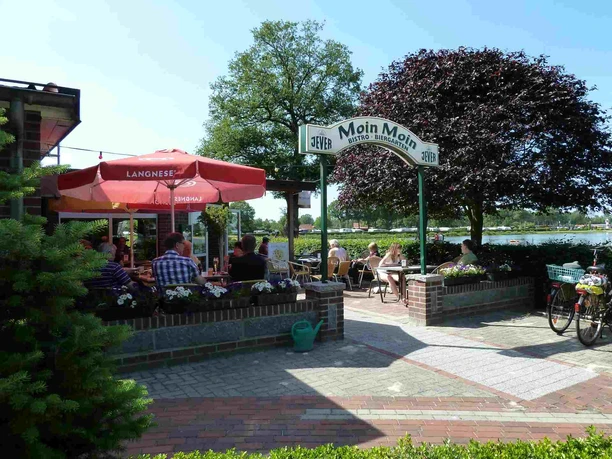 Guests sit under a red parasol in front of the "Bistro Moin Moin". An inviting entrance with flowers leads into the cozy café. Bicycles are parked in the foreground, a beautiful day by the water.