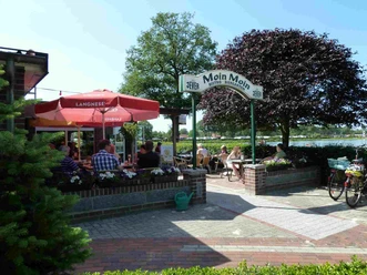 Bistro Moin Moin Vor dem "Bistro Moin Moin" sitzen Gäste unter einem roten Sonnenschirm. Ein einladender Eingang mit Blumen führt in das gemütliche Café. Fahrräder sind im Vordergrund abgestellt, ein schöner Tag am Wasser.