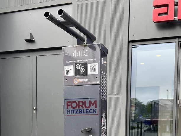 Fahrrad-Reparaturstation Parkplatz Forum Hitzbleck Fahrrad-Reparaturstation mit Werkzeugen und Luftpumpe auf dem Parkplatz Forum Hitzbleck. Symbole für Fahrrad, Kinderwagen und Rollstuhl sind sichtbar.