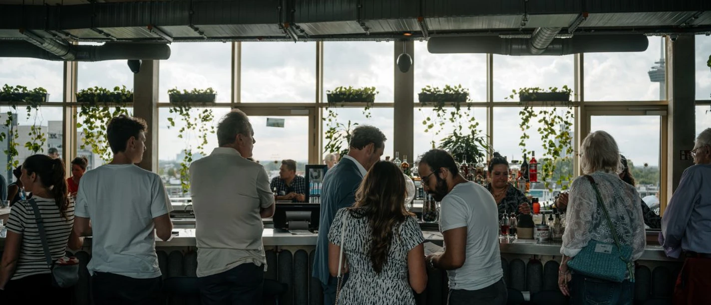 Monkey Bar Innenbereich Menschen stehen an einer Bar vor großen Fenstern mit Pflanzen und lichtdurchflutetem Raum.