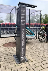 Fahrrad-Reparaturstation Parkplatz Forum Hitzbleck Fahrrad-Reparaturstation mit Werkzeugen, ein Fahrrad mit Anhänger und Einkaufswagen im Hintergrund.