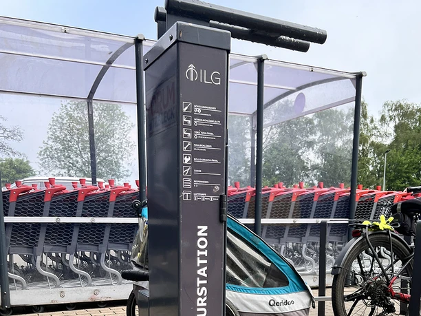 Fahrrad-Reparaturstation Parkplatz Forum Hitzbleck Fahrrad-Reparaturstation mit Werkzeugen, ein Fahrrad mit Anhänger und Einkaufswagen im Hintergrund.