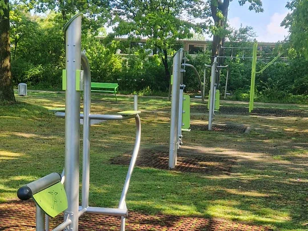 Outdoorfitness-Anlage Vorwerk Outdoor-Fitnessgeräte aus silbernem Metall mit grünen Akzenten in einem Park mit hohen Bäumen.
