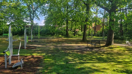 Bild-Standort-Vorwerk-Querformat.jpg Eine Outdoorfitness-Anlage mit verschiedenen Sportgeräten auf einer Wiese unter grünen Bäumen.