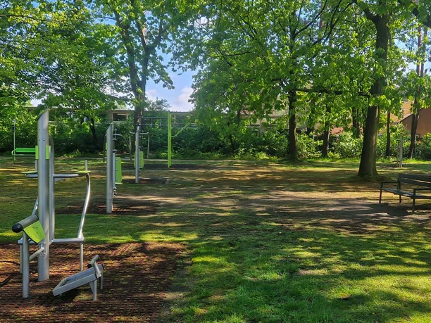 Bild-Standort-Vorwerk-Querformat.jpg Eine Outdoorfitness-Anlage mit verschiedenen Sportgeräten auf einer Wiese unter grünen Bäumen.