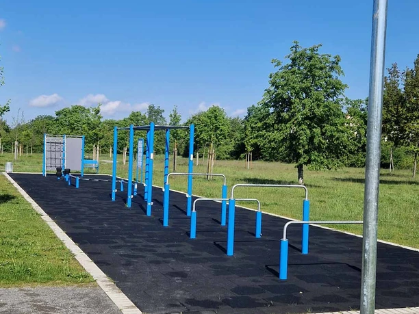 Outdooranlage-Altencelle Ein Outdoor-Fitnesspark mit blauen Geräten auf einer schwarzen Bodenmatte in einer grünen Parkanlage.