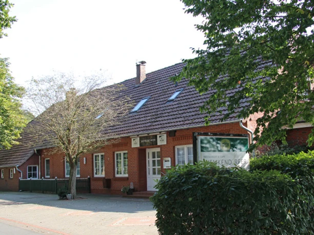 Außenansicht Ein rotes Backsteingebäude mit vielen Fenstern unter einem Schrägdach, umgeben von Bäumen.